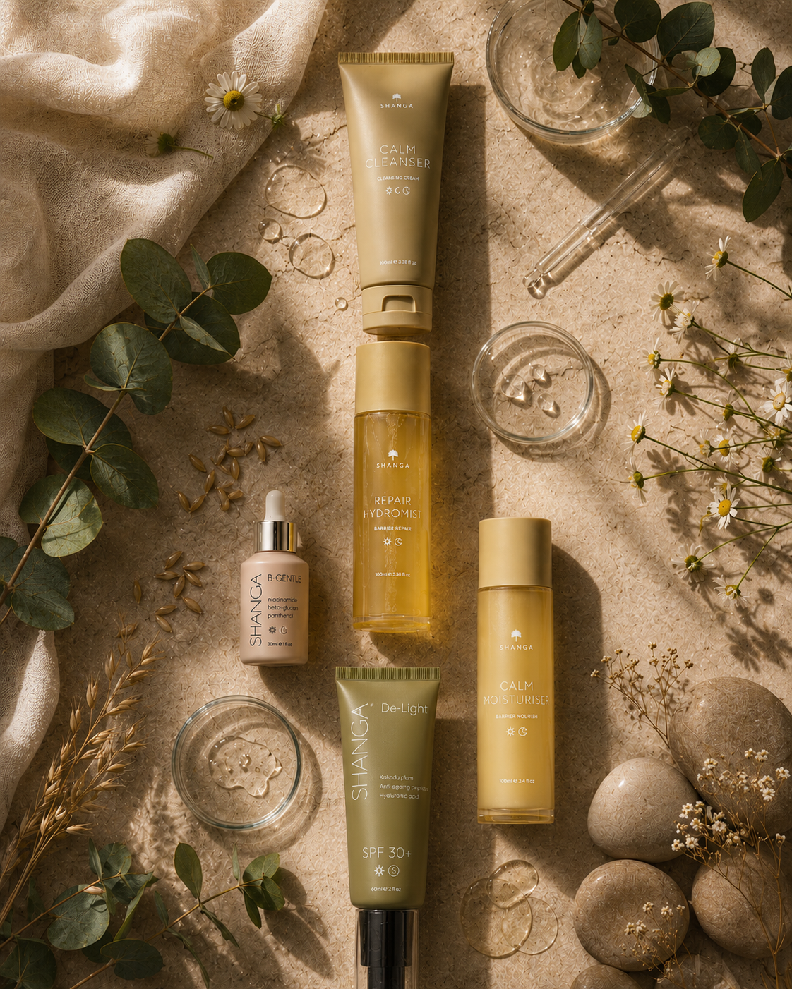 Five Shanga skincare products arranged in a styled flat lay on a textured beige surface with eucalyptus leaves, dried flowers, glass droppers, and stones.